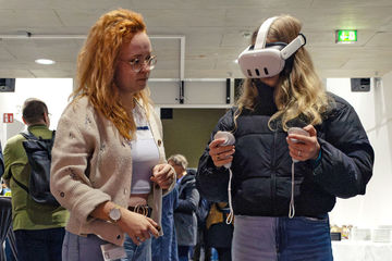 Zwei Frauen stehen nebeneinander, eine trägt eine VR-Brille und hält entsprechende Controller in der Hand.