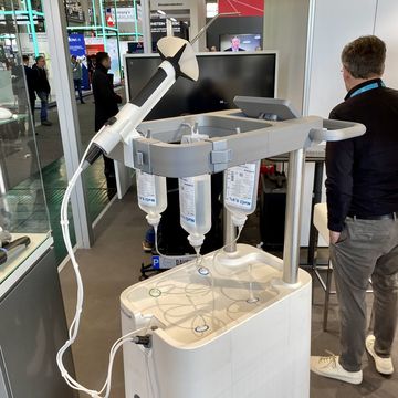 An einem fahrbaren Gestell hängen Infusionsbeutel. Mit einem Schlauch ist zudem ein Handgerät verbunden, das aus einem langen Stab, einem Trichter und einem langen dünnen Rohr besteht.