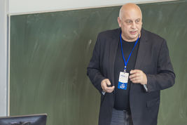 Zu sehen ist ein Mann vor eine Wandtafel, der gestikulierend eine Rede hält.