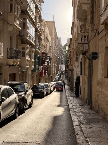 Das Bild zeigt eine Straße, die sich zwischen zwei Häuserwänden erstreckt und auf der Fahrzeuge und Menschen zu sehen sind.