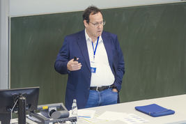 Zu sehen ist ein Mann vor eine Wandtafel, der eine Rede hält und dabei mit einer Hand gestikuliert.