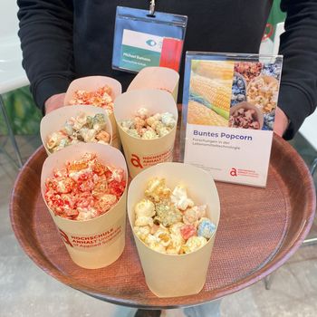 Zu sehen ist ein Tablett, das von einer Person gehalten wird und auf dem mehrere Becher stehen, die bunt eingefärbtes Popcorn beinhalten.
