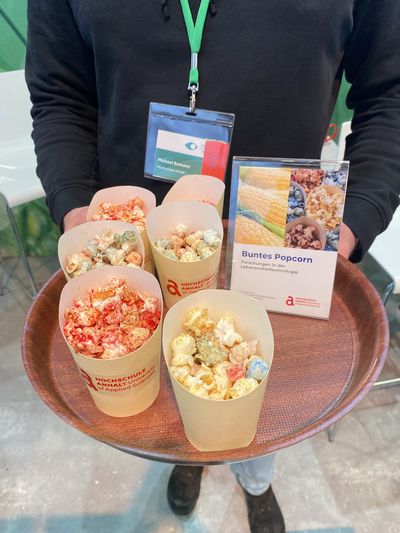 Zu sehen ist ein Tablett, das von einer Person gehalten wird und auf dem mehrere Becher stehen, die bunt eingefärbtes Popcorn beinhalten.