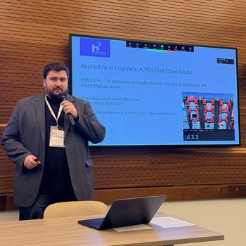 Das Bild zeigt einen Referenten mit Mikrofon, der auf dem Tisch vor sich einen offenen Laptop stehen hat. Im Hintergrund ist ein Bildschirm zu sehen, der eine Vortragsfolie zeigt.