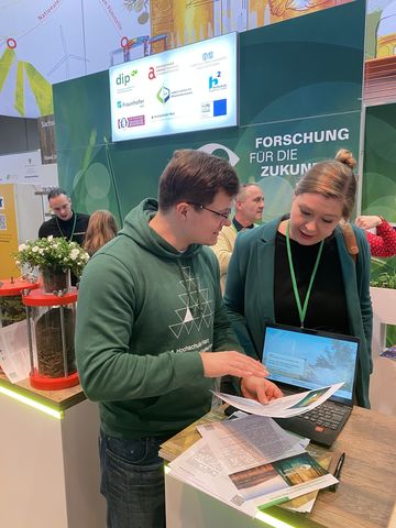 An einem Messestand befinden sich zwei Personen im Gespräch und blicken dabei auf ein Schriftstück.