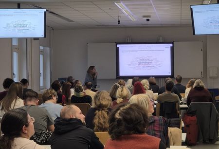 Zahlreiche Personen sind von hinten sitzend zu sehen und folgen den Ausführungen einer Person, die Inhalte auf einem Bildschirm präsentiert.