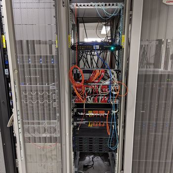 Es ist ein schmaler, hoher Metallschrank zu sehen, in dem sehr viele verschiedenfarbige LAN-Kabel angeschlossen sind.