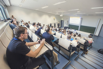 Es ist ein Hörsaal zu sehen, in dem zahlreiche Personen auf den Plätzen sitzend in Richtung einer Frau blicken, die eine Rede hält. Dahinter zu sehen ist eine Präsentationsfolie, die auf die Wand projiziert wird.