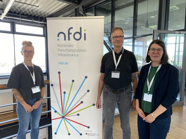 Zwei Frauen und ein Mann stehen neben einem Roll-up-Banner, auf welchem das Logo des NFDI abgebildet ist.