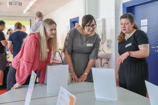 Drei Frauen schauchen sich interessiert Poster an, die in einem Tischaufsteller präsentiert werden.