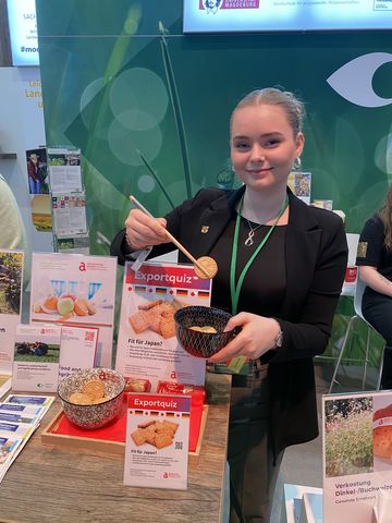 Eine Frau steht hinter einem Messestand auf dem Aufsteller stehen und hält einen Keks mit Essstäbchen fest.
