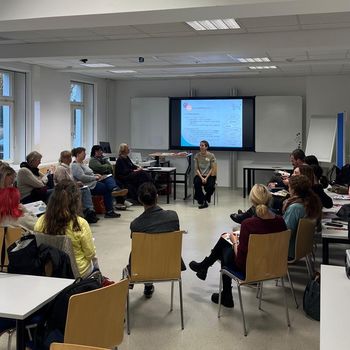 Zu sehen ist ein Konferenzraum, in dem mehrere Personen auf im Kreis angeordneten Stühlen sitzen und miteinander diskutieren.