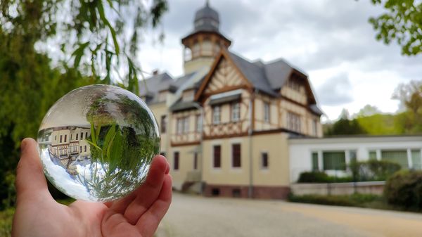 Das Bild zeigt im Hintergrund eine Fachwerkvilla. Im Vordergrund ist eine Hand mit einer Glaskugel zu sehen, die das Gebäude auf dem Kopf stehend widerspiegelt.