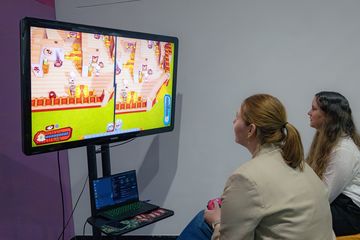 Zwei junge Frauen sitzen vor einem großen Monitor. Sie halten Spielecontroller in der Hand. Auf dem Bildschirm ist die Grafik eines Videospiels zu erkennen.