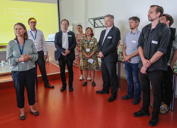 Mehrere Personen stehen nebeneinander. Sie hören einer Ansprache zu. Im Hintergrund ist eine Leinwand mit einer Präsentationsfolie zu sehen.