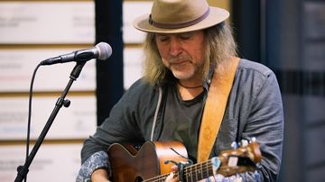 Ein Mann steht an einem Mikrophon. Er trägt zudem eine Gitarre und spielt darauf.
