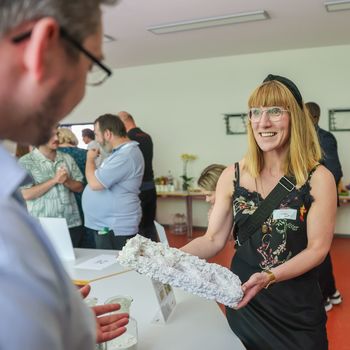 Eine Frau hält ein weißes, steinartiges Gebilde in der Hand. Sie lächelt. Ein Mann schaut interessiert auf das Objekt. Im Hintergrund sind weitere Menschen zu sehen, die sich unterhalten.