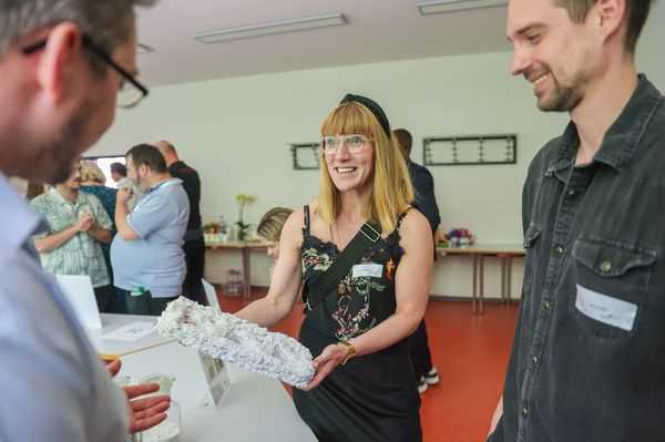 Eine Frau hält ein weißes, steinartiges Gebilde in der Hand. Sie lächelt. Zwei Männer schauen interessiert auf das Objekt. Im Hintergrund sind weitere Menschen zu sehen, die sich unterhalten.